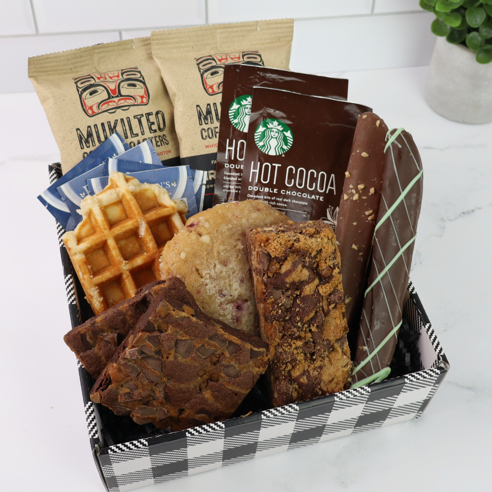 Assorted baked goods gift box including waffles, cookies, coffee, tea, brownies and hot cocoa mix in a box on a white surface.